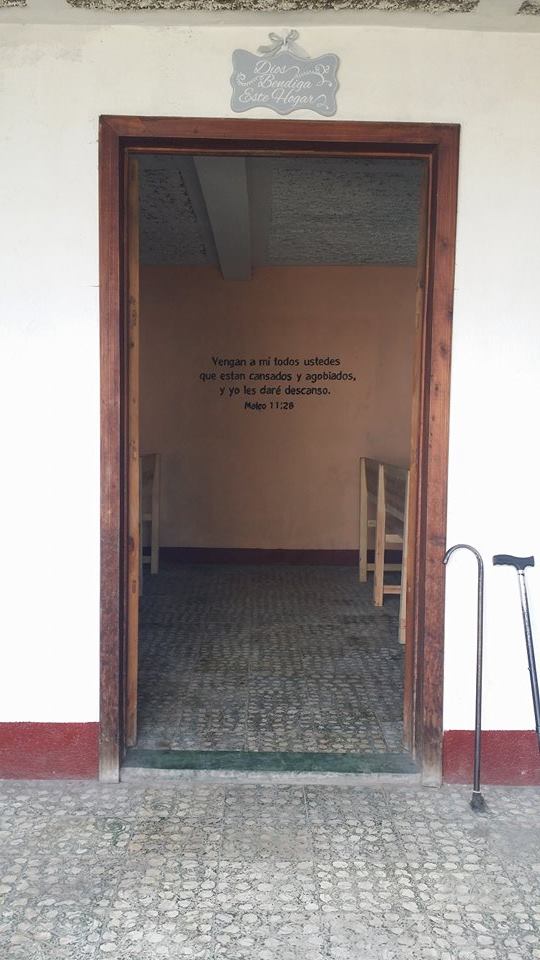 The door to the dining room with canes propped outside, a “God Bless this Home” sign above the door in Spanish, and Matthew 11:28 on the wall.